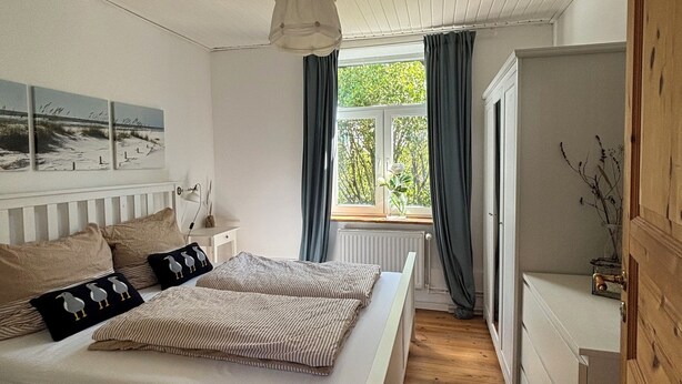 A bedroom with a large bed, a window with curtains, a wooden floor, and a view of greenery outside. - Cozy Scandinavian-style vacation home between the Schlei and the Baltic Sea (Rieseby)