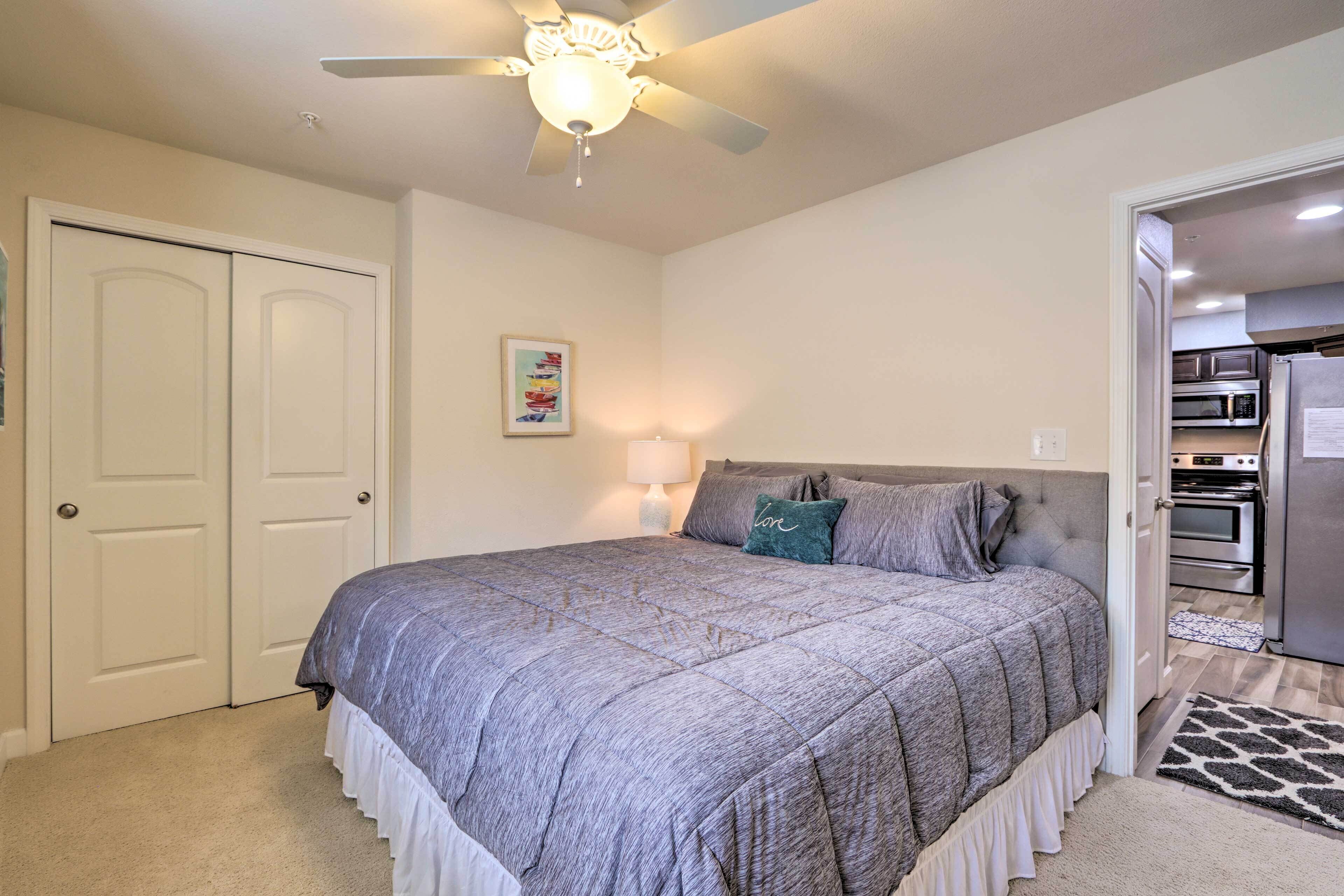 Ein Schlafzimmer mit Bett, Deckenventilator, Tür und Blick zur Küche.