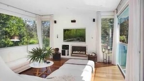 A modern living room with a sofa, a coffee table, and a TV. There is a large window with a view of trees. - Colenso Luxury Home, Perfect for Friends & Family (Erina)