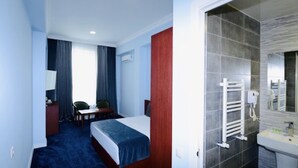 Une chambre d’hôtel comprenant un lit, un bureau, une chaise, une petite table, une lampe et une salle de bain avec un lavabo et un miroir.