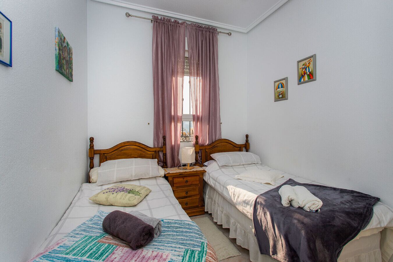 Een slaapkamer met twee eenpersoonsbedden, een houten hoofdbord, een nachtkastje, een raam met gordijnen en wanddecoratie.
