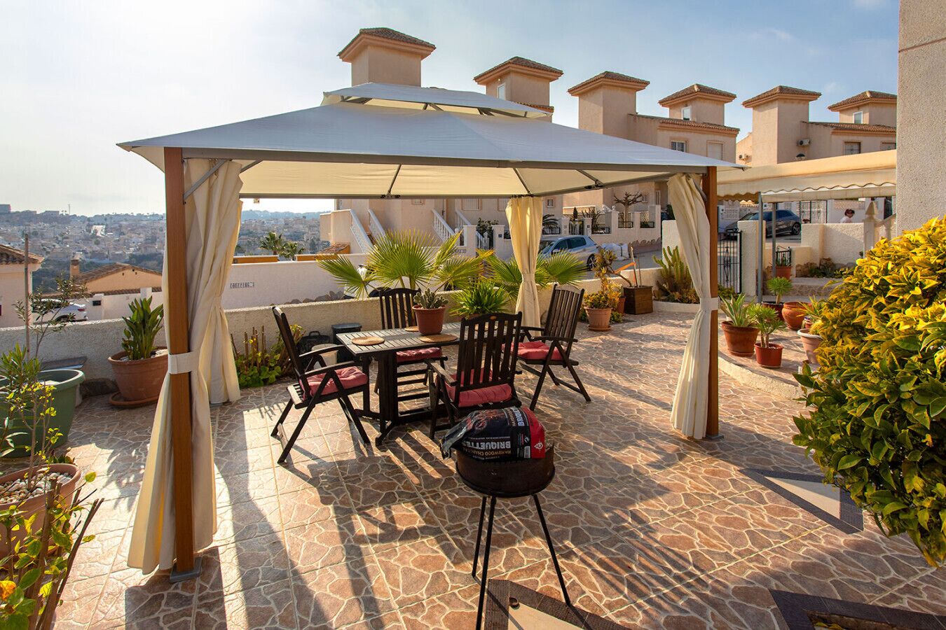 A covered outdoor patio with a table and chairs, potted plants, and a view of the city.