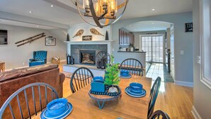 Un comedor con una mesa de madera, platos azules y sillas. Hay una chimenea con repisa, un sillón azul y un televisor en la pared.