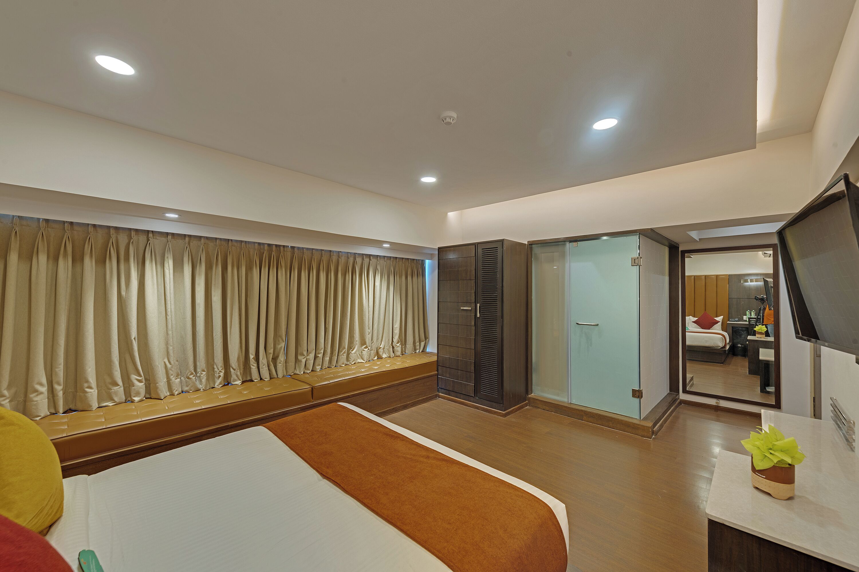 A hotel room with a bed, a seating area, a wardrobe, a television, and a bathroom visible through a glass door.