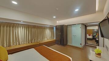 A hotel room with a bed, a seating area, a wardrobe, a television, and a bathroom visible through a glass door.