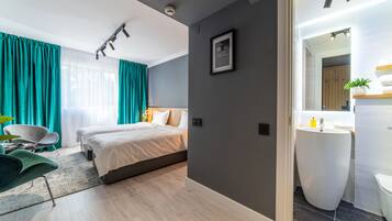 Una habitación de hotel moderna con una cama grande, una zona de estar con una silla, y un baño con lavamanos y espejo.