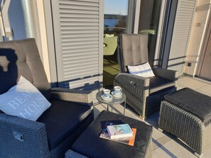 Varanda com mobiliário de vime, incluindo um sofá e cadeiras, uma mesa pequena com um livro e uma xícara, e vista para o mar.