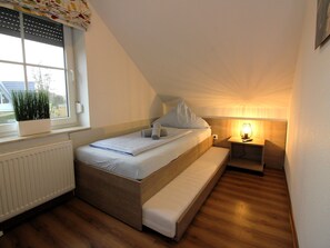 A small, cozy bedroom with a single bed, a bench, a nightstand with a lamp, and a window with blinds. - ROB 01 for 6 persons, approx. 78 sqm - Feriendorf Robbenplatte Burhave (1) (Butjadingen)