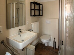 A bathroom with a white sink, a toilet, a glass shower enclosure, a radiator, and a small cabinet above the toilet. - ROB 01 for 6 persons, approx. 78 sqm - Feriendorf Robbenplatte Burhave (1) (Butjadingen)