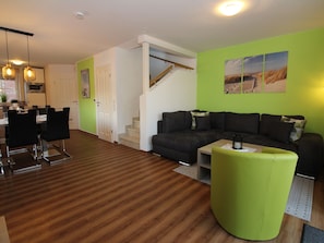 Uma sala de estar moderna com um sofá escuro, um banquinho verde e um espaço de jantar com cadeiras e uma mesa.