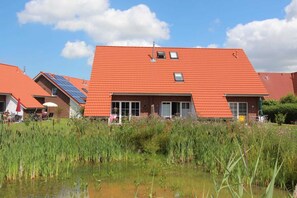 A building with a red-tiled roof, solar panels, and a pond in the foreground. - ROB 01 for 6 persons, approx. 78 sqm - Feriendorf Robbenplatte Burhave (1) (Butjadingen)