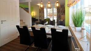 Un comedor con una mesa lista para cuatro personas, sillas negras, lámparas colgantes y vistas al exterior a través de una ventana con cortinas.