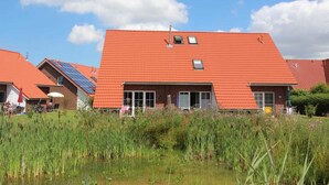 A building with a red-tiled roof, solar panels, and a pond in the foreground. - ROB 02 for 6 persons, approx. 78 sqm - Feriendorf Robbenplatte Burhave (1) (Butjadingen)