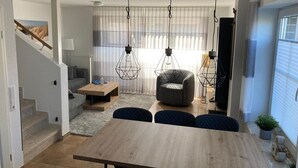 Un espace de vie moderne avec une table à manger en bois, un fauteuil gris et un canapé. On y trouve des luminaires suspendus et un escalier menant à un niveau supérieur.