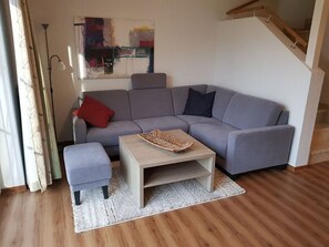 Un salon moderne avec un canapé d’angle gris, un petit pouf et une table basse en bois clair. Un grand tableau abstrait orne le mur, et un escalier se trouve au fond de la pièce.