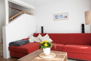 Um sofá vermelho com uma almofada branca, uma mesa de centro de madeira com um vaso branco de flores amarelas e um quadro na parede.