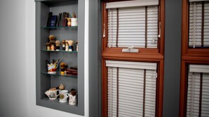 Una stanza con una parete grigia, una finestra con cornice in legno e persiane, e uno scaffale con diversi oggetti, tra cui libri, pennelli e decorazioni.