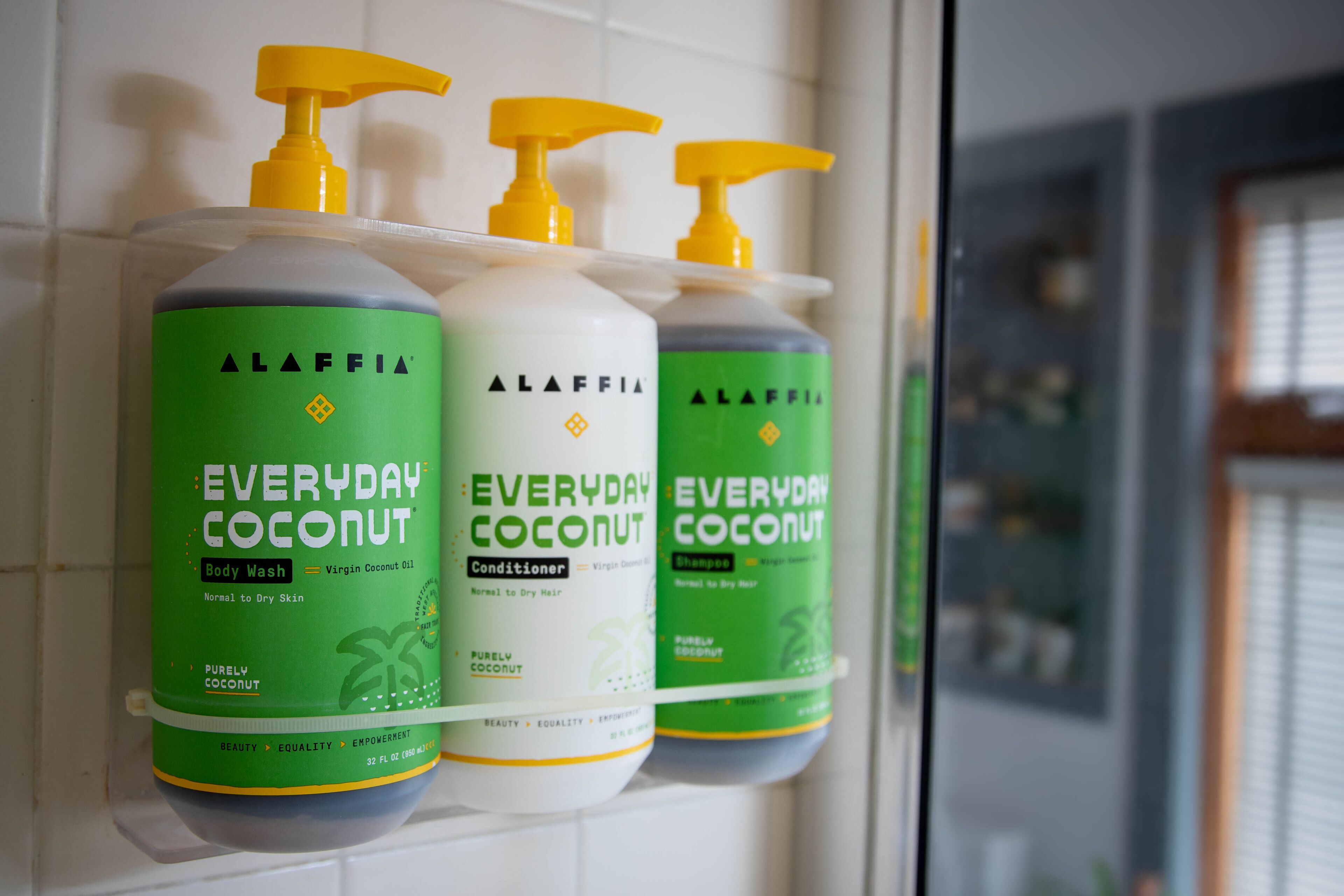 Three bottles of Alaffia Everyday Coconut body wash, conditioner, and shampoo on a white tiled wall.