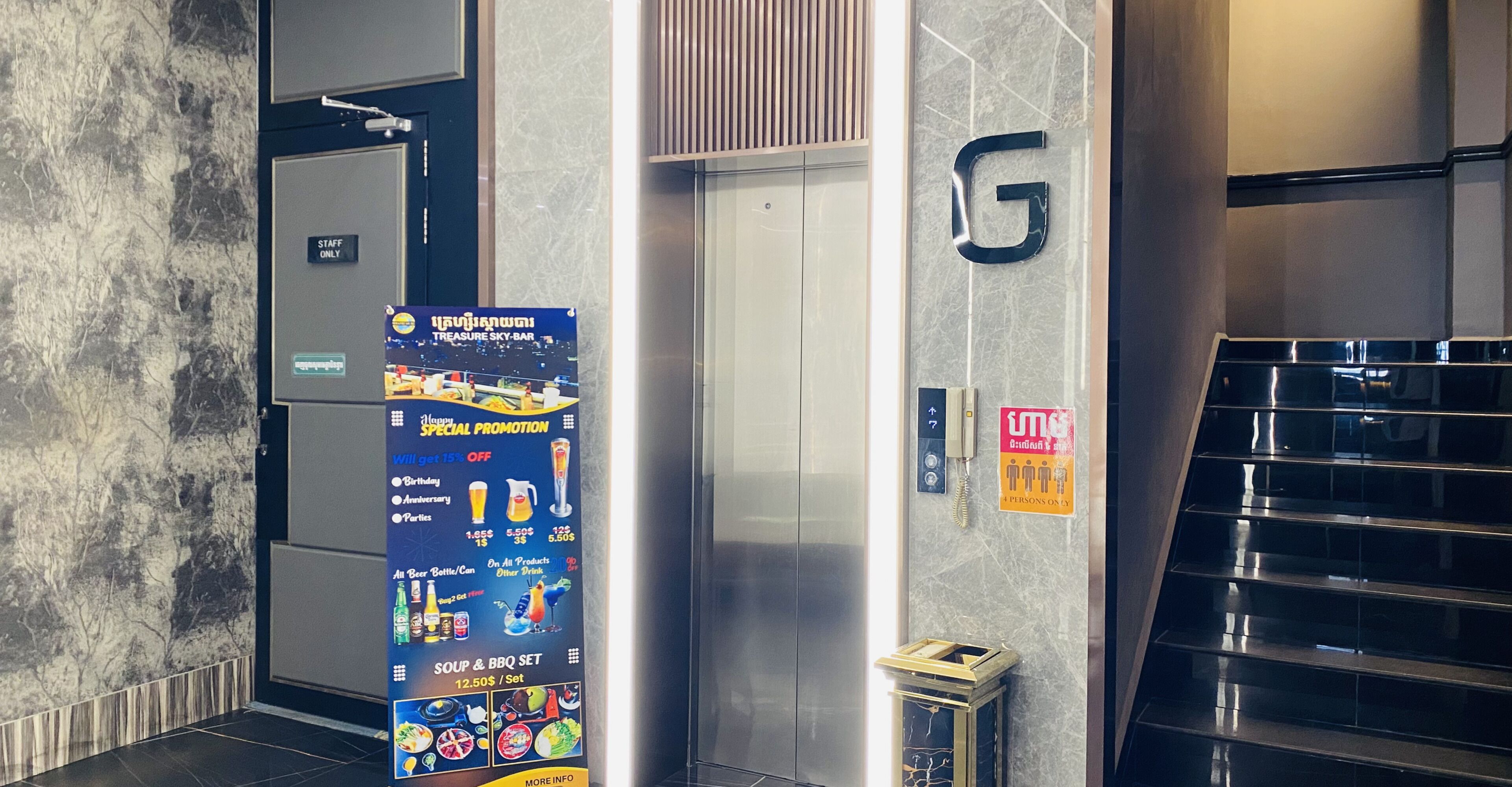 Een liftinterieur met een 'Staff Only' bord, een reclameposter voor 'Treasure Sky-Bar', een 'G' bord en een telefoon.