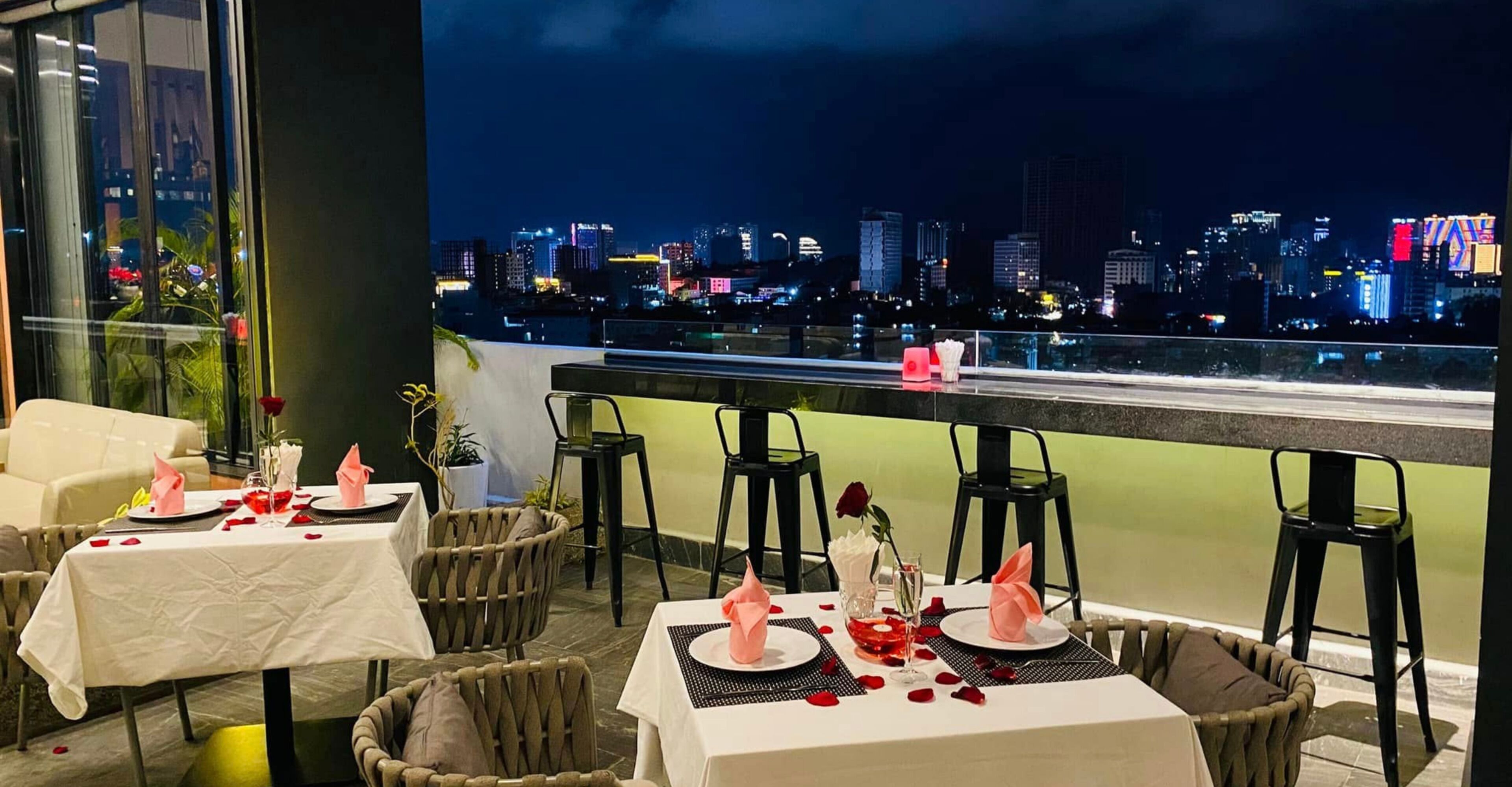 Un espacio de comedor en la azotea con mesas dispuestas para dos, y vistas panorámicas del horizonte urbano.
