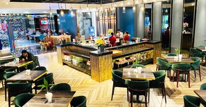 Un restaurante con sillas tapizadas de verde, mesas de madera y una barra central con personal.