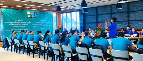 A meeting room with attendees seated at tables, a banner on the wall, and a person standing addressing the group.