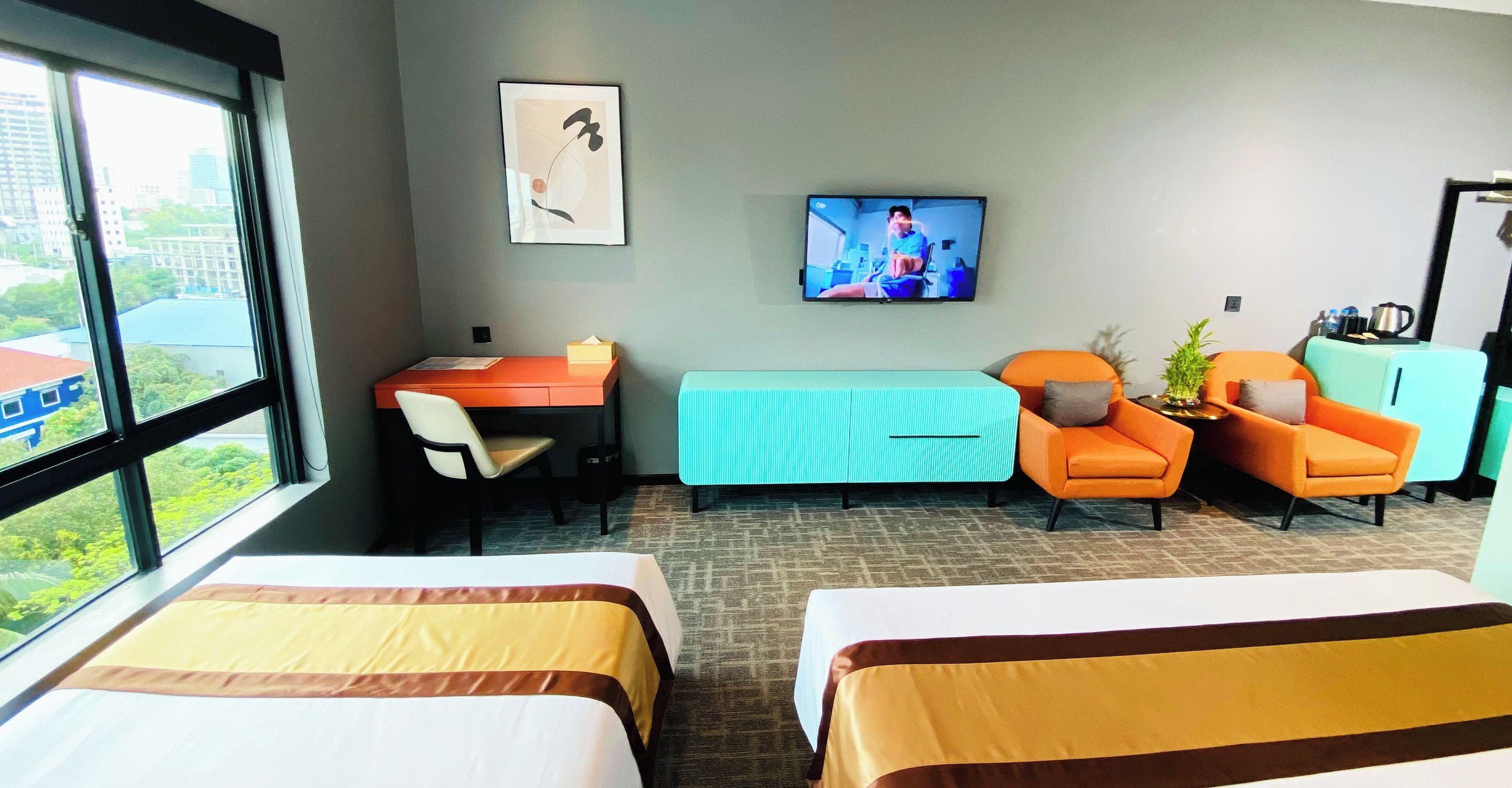 Une chambre d’hôtel avec deux lits, un bureau, un fauteuil orange, un canapé turquoise, une télévision et une fenêtre donnant sur la ville.
