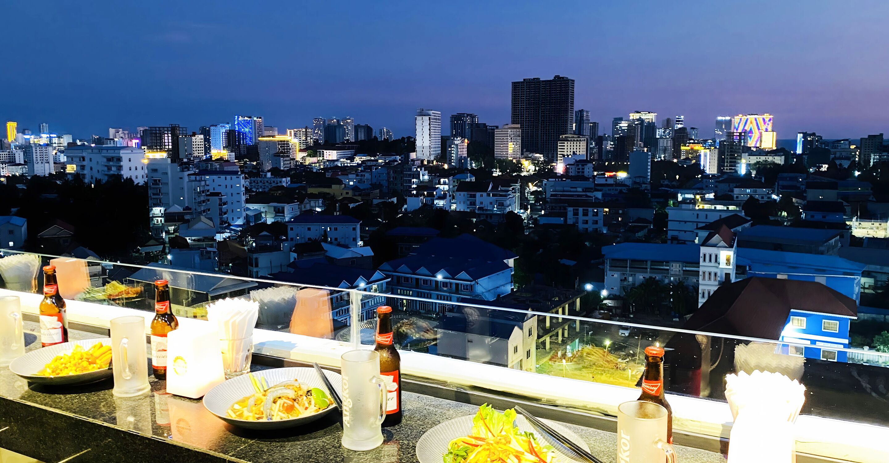 A rooftop dining area with a cityscape view, tables set with plates of food, and bottles of beer.