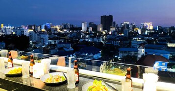 Un área de comedor en la azotea con vistas a la ciudad, mesas dispuestas con platos de comida y botellas de cerveza.