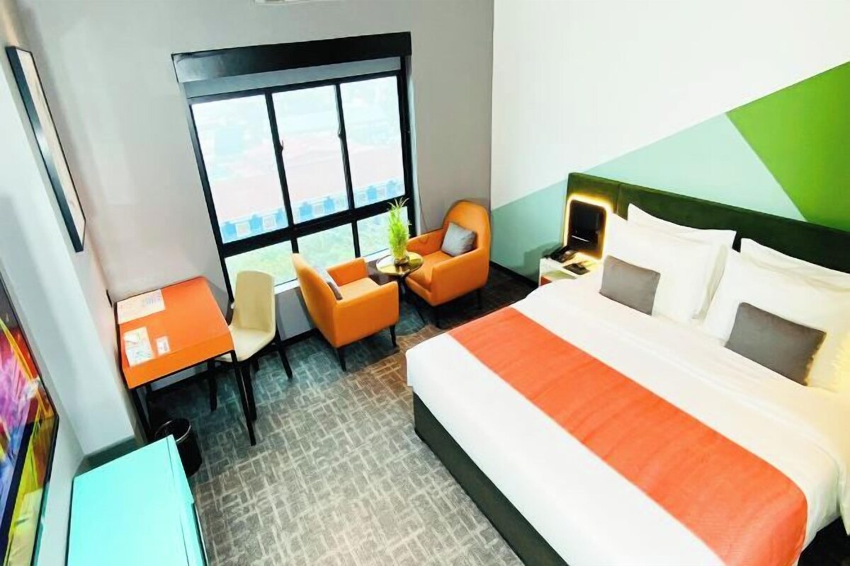 Une chambre d’hôtel avec un grand lit, un bureau avec une chaise, une petite table avec une plante et une fenêtre donnant sur l’extérieur.