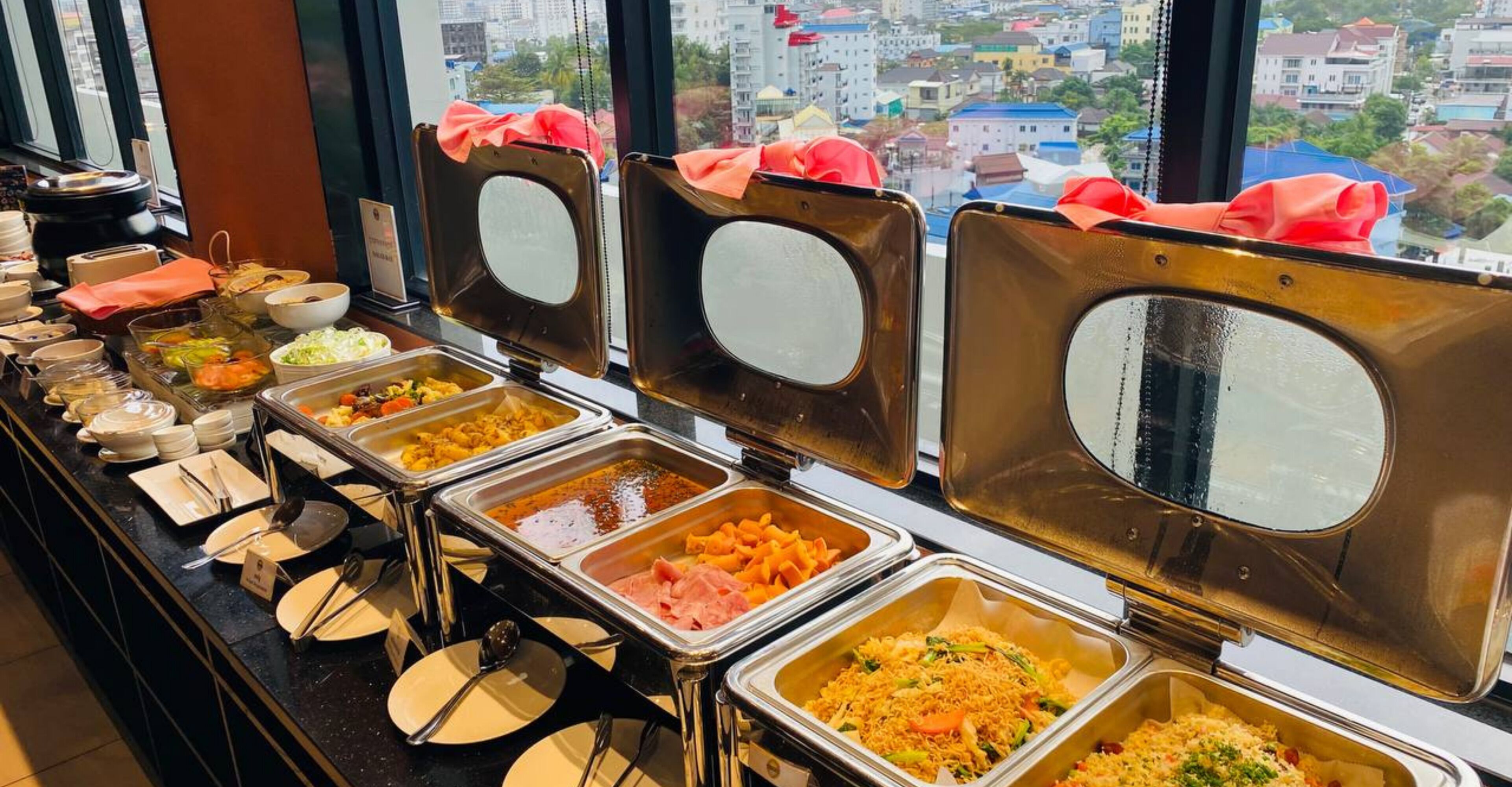A buffet with various food options and a city view through the window.