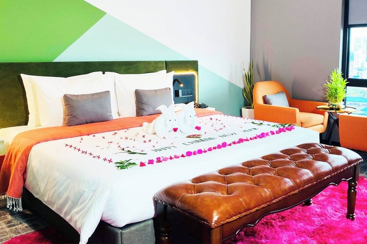 Habitación de hotel con cama, una banca, una silla, una mesita y ventana con vista a la ciudad.