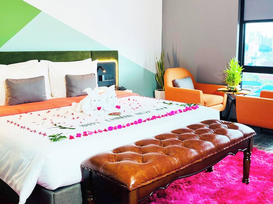 Habitación de hotel con cama, una banca, una silla, una mesita y ventana con vista a la ciudad.