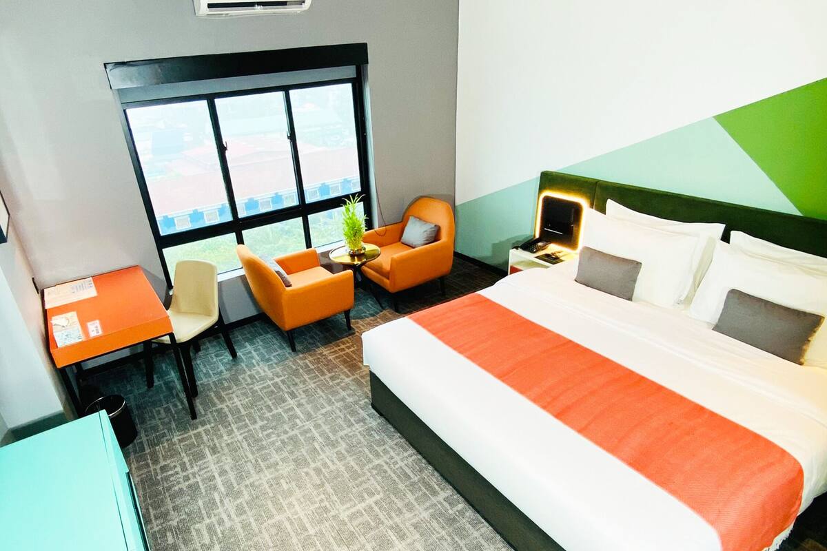 Habitación de hotel con una cama grande, un escritorio, dos sillas, una mesita con una planta y una ventana con vistas.