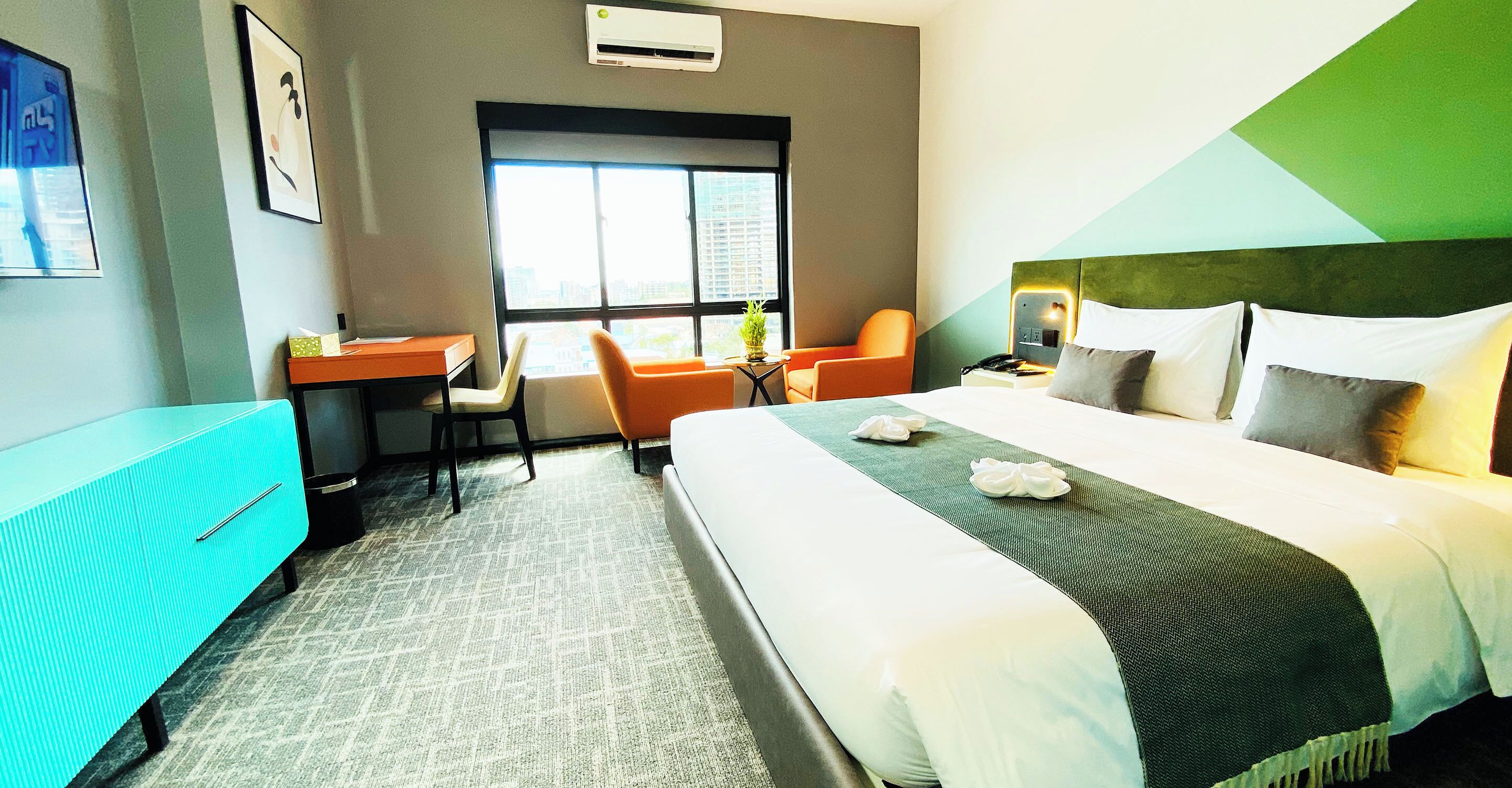 A hotel room with a bed, a desk, two chairs, a window, and a green accent wall.