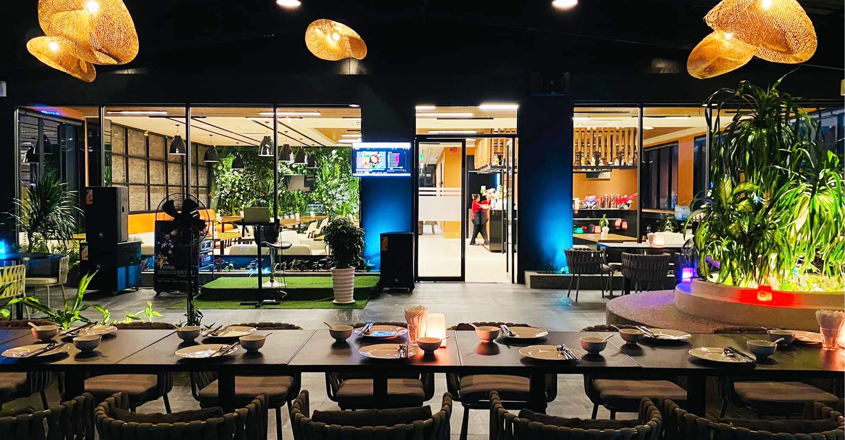 A restaurant with tables set for dining, a digital menu display, and indoor plants.