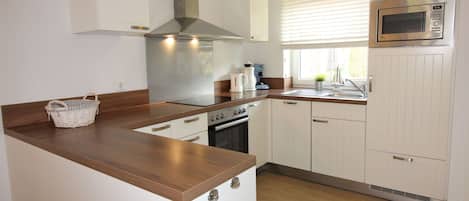 Een praktische keuken met een centraal eiland, witte keukenkastjes, een roestvrijstalen afzuigkap, een magnetron en een raam met jaloezieën.