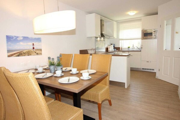 A dining area with a wooden table set for four, surrounded by wicker chairs. A large white lamp hangs above the table. The kitchen is visible in the background with white cabinets and a microwave.