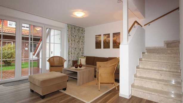 A spacious living room with a sofa, ottoman, and chairs, a staircase, and large windows with a view of a brick building and greenery. - ROB 15 for 8 persons, approx. 86 sqm - Feriendorf Robbenplatte Burhave (2) (Butjadingen)