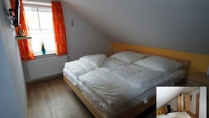 Een kleine eenpersoonskamer met een schuine plafond, een raam met oranje gordijnen, een radiator en een klein plankje met een plantje.