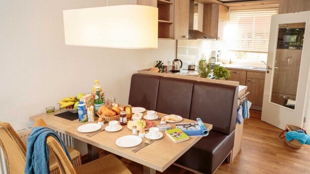 Een gezellige eethoek met een houten tafel gedekt voor het ontbijt, een bruine leren bank en een keuken op de achtergrond.
