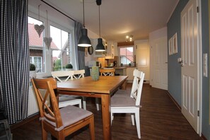 Un coin repas avec une table en bois et des chaises blanches, une cuisine en arrière-plan et, à l’extérieur, une vue sur un bâtiment au toit rouge.