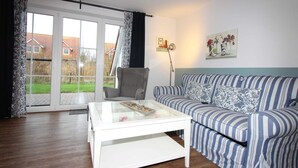 A living room with a striped sofa, a glass coffee table, a grey armchair, and a large window with curtains. - ROB 20 for 8 persons, approx. 86 sqm, pets, sauna - Feriendorf Robbenplatte (Butjadingen)