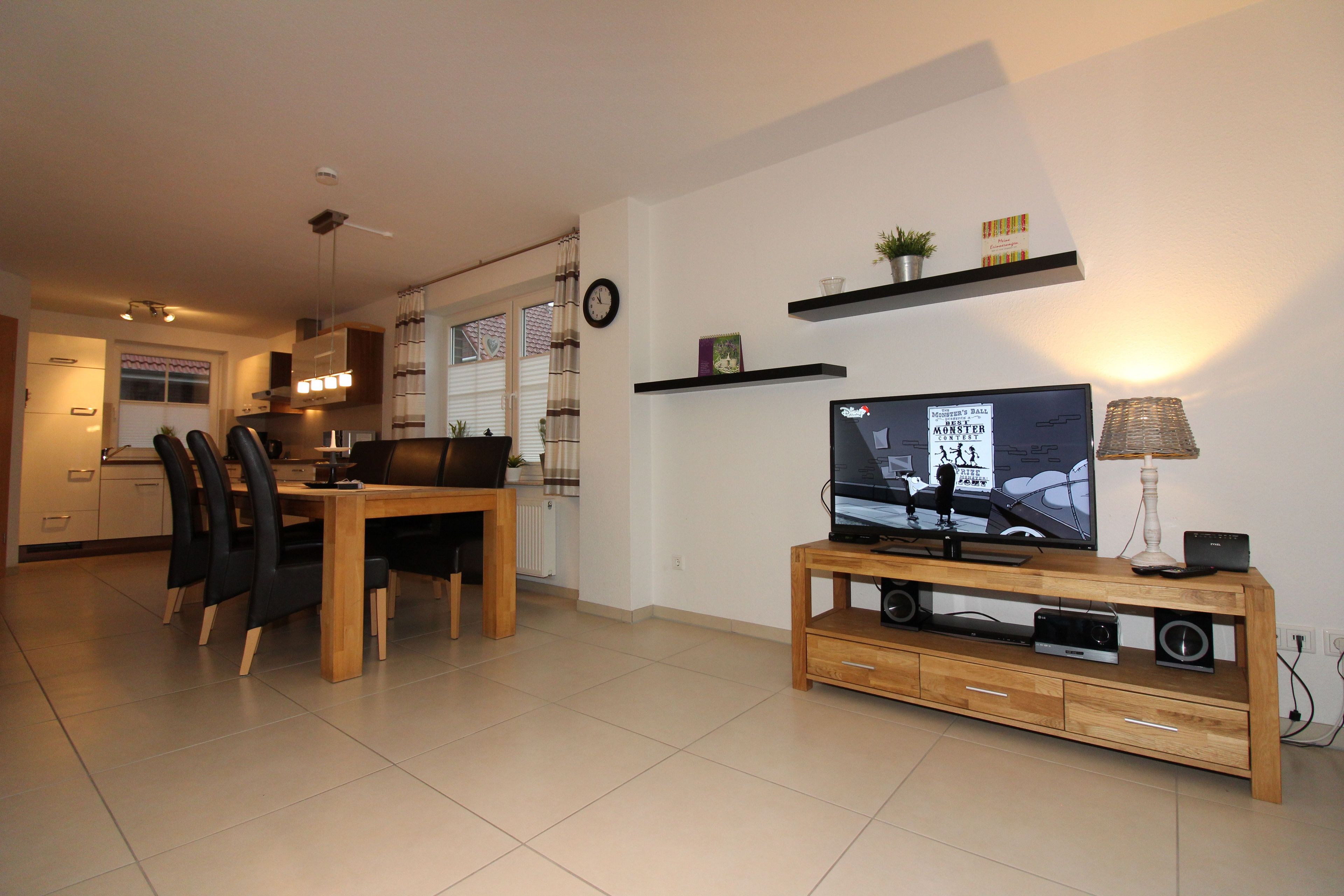A modern living room with a television on a wooden stand, a dining area with a table and chairs, and a kitchen in the background.