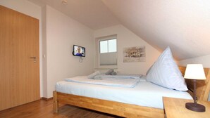 A bedroom with a wooden bed, a nightstand with a lamp, a window with blinds, a door, and a TV on the wall. - ROB 18 for 6 persons, approx. 86 sqm - Feriendorf Robbenplatte Burhave (3) (Butjadingen)