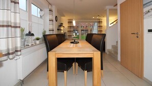 A dining area with a wooden table and black chairs, a radiator, and a view of a residential building through large windows. - ROB 18 for 6 persons, approx. 86 sqm - Feriendorf Robbenplatte Burhave (3) (Butjadingen)