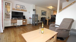 A modern living room with a flat-screen TV, a wooden cabinet, a fireplace, a dining area with a table and chairs, and a comfortable armchair. - ROB 19 for 6 persons, approx. 86 sqm, sauna - Feriendorf Robbenplatte Burhave (3) (Butjadingen)