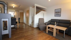 A modern living room with a sofa, a coffee table, and a fireplace. There is a staircase leading to another level, and a dining area with a table and chairs. - ROB 19 for 6 persons, approx. 86 sqm, sauna - Feriendorf Robbenplatte Burhave (3) (Butjadingen)