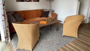 A living room with a sofa, two wicker chairs, a coffee table, and a lamp. There are two wall-mounted pictures depicting a beach scene. - ROB 16 for 7 persons, approx. 86 sqm - Feriendorf Robbenplatte Burhave (3) (Butjadingen)