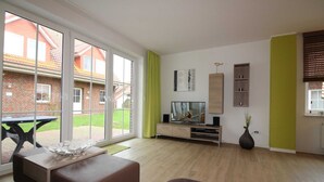 A modern living room with a large sliding glass door leading to a patio, a flat-screen TV, a wooden cabinet, and a vase with flowers. - ROB 17 for 6 persons, approx. 86 sqm, pets - Feriendorf Robbenplatte Burhave (Butjadingen)